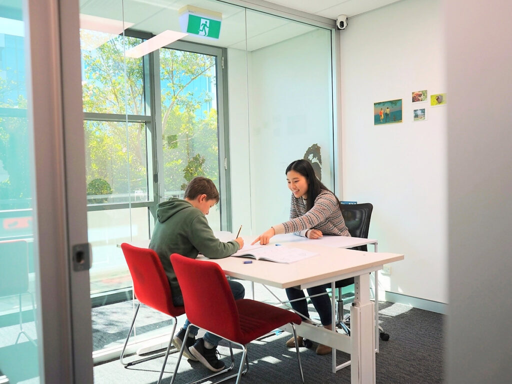Programs A tutor helping a primary school student with a maths workbook at a white desk